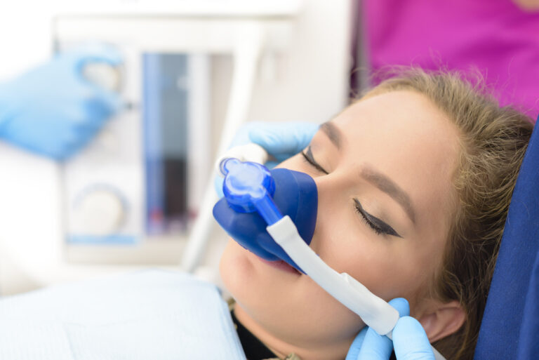 A patient lies in a dental chair wearing a nasal sedation mask while a clinician assists. The image represents dental sedation used to help patients relax during treatment.